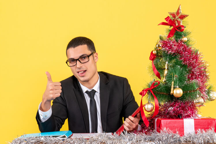 男人正面图：穿着西装的快乐的年轻人坐在桌子旁做着竖起大拇指的圣诞树和礼物商人桌子年轻人