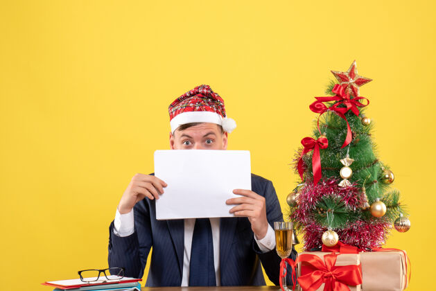 好奇的人神秘人的前视图 他用文件盖住脸 坐在圣诞树旁的桌子上 黄色的礼物脸礼物桌子