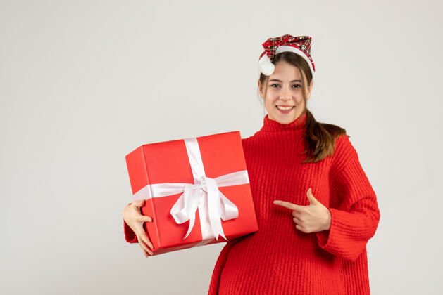 礼物戴着圣诞帽的快乐女孩拿着礼物手指着她的礼物站在白色的地板上圣诞老人拿着站着