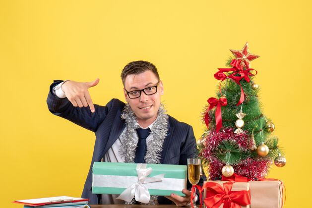 圣诞正面图：快乐的男人指着坐在圣诞树旁桌上的礼物和黄色的礼物礼物快乐男人坐着