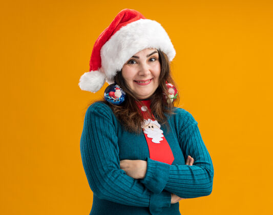 帽子微笑的成年白人妇女戴着圣诞帽 打着圣诞领带 耳朵上戴着玻璃球饰物 交叉双臂站在橙色背景上 留有复制空间微笑胳膊圣诞老人