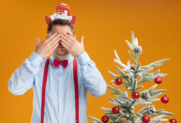 盖戴着吊带领结的年轻人站在圣诞树旁 戴着圣诞老人和红眼镜 用手捂住橙色的墙站立男人吊带