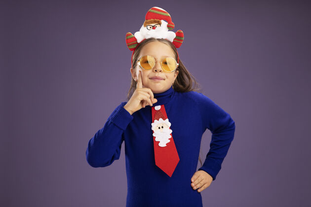 滑稽小女孩穿着蓝色高领毛衣 系着红色领带 头上戴着有趣的圣诞戒指 站在紫色的背景下 面带微笑地看着相机头高领毛衣边缘