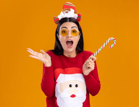 头带令人印象深刻的年轻白人女孩戴着圣诞老人的头带和毛衣与眼镜举行传统的圣诞糖果手杖显示空手孤立在橙色的墙上空圣诞老人毛衣