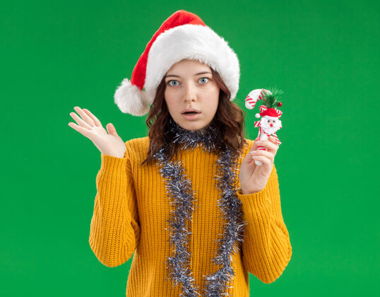 圣诞老人震惊的年轻斯拉夫女孩 戴着圣诞帽 脖子上戴着花环 手里拿着糖果手杖 手一直张开手花环糖果