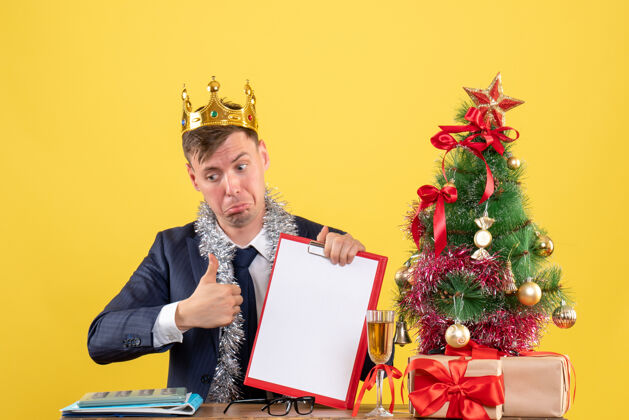 树商务人士在圣诞树旁的桌子旁做竖起大拇指的招牌 黄色的礼物免费商务男性