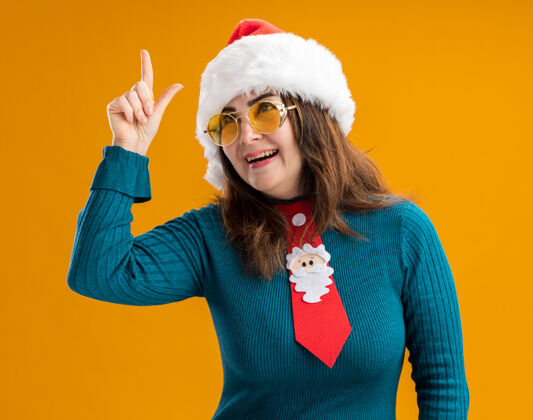 成人快乐的成年白种女人戴着太阳眼镜 戴着圣诞帽 系着圣诞领带 在橙色的背景上孤立地指向上 还有复制空间快乐帽子眼镜