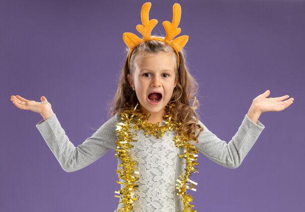 穿兴奋的小女孩戴着圣诞发箍 脖子上戴着花环 双手分开放在蓝色的墙上圣诞节蔓延小