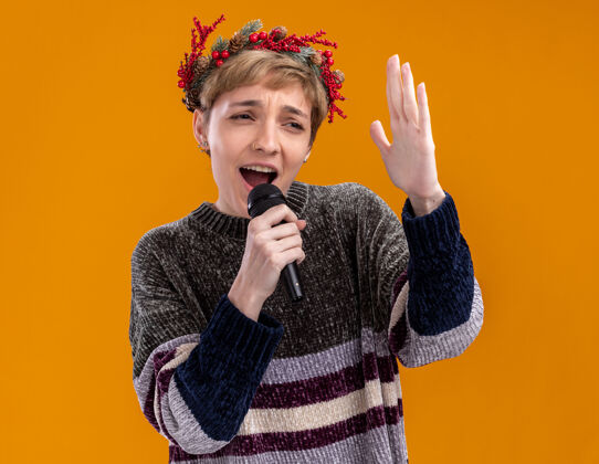 圣诞节年轻漂亮的女孩戴着圣诞花环 拿着麦克风近嘴 手举在空中 看着边唱隔离在橙色墙上穿着麦克风头