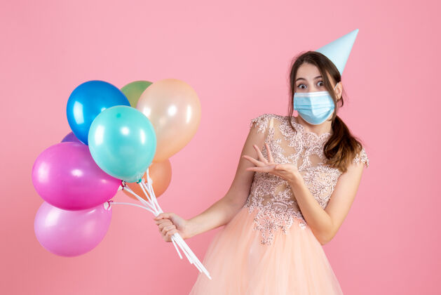 帽子一个戴着派对帽和医用面具的惊喜女孩 手里拿着粉色的彩色气球生日气球举行