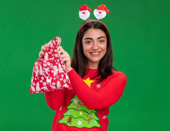 包微笑着的年轻白人女孩 戴着圣诞老人的头带 手里拿着圣诞礼品袋 绿色背景上有复制空间圣诞老人圣诞节女孩