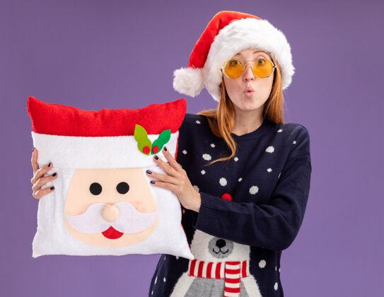 帽子关心的年轻漂亮女孩穿着圣诞毛衣戴着圣诞帽戴着眼镜抱着圣诞枕头孤立在紫色的墙上圣诞节枕头穿着