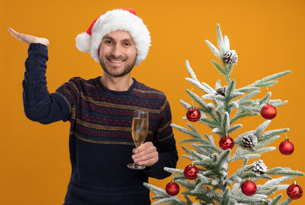 站着快乐的白人年轻人戴着圣诞帽 站在装饰好的圣诞树旁 手里拿着一杯香槟 孤零零地站在橙色的墙上戴着空着靠近
