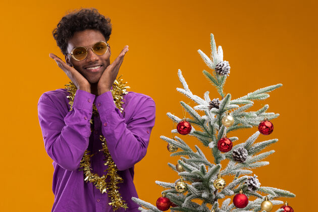 周围兴奋的非洲裔美国年轻人戴着眼镜 脖子上戴着金箔花环 站在装饰过的圣诞树旁 手放在脸上 隔离在橙色的墙上脖子金箔男人
