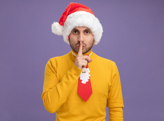 领带令人印象深刻的年轻白种人戴着圣诞帽 打着领带 做着沉默的手势 孤立地在紫色的墙上复制空间手势男人帽子