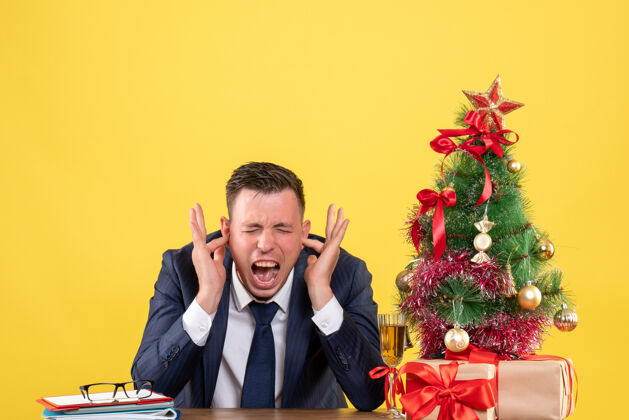 商人前视图中的人大声喊叫 手指在耳朵里 坐在圣诞树旁的桌子上 黄色的礼物耳朵男人西装