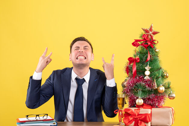 黄色圣诞树旁坐在桌旁的欢欣男子的正面图 黄色的礼物坐着办公室树