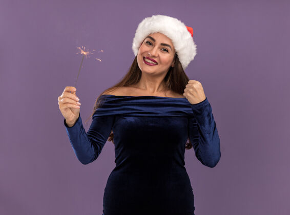 帽子微笑着的年轻漂亮女孩穿着蓝色连衣裙 戴着圣诞帽 手里拿着闪闪发光的灯 在紫色的背景上显示“是”的手势手势裙子是的