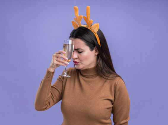 抱着累得闭着眼睛年轻漂亮的女孩穿着棕色毛衣圣诞发箍捧着香槟在额头上蓝色背景隔离香槟圣诞节头发