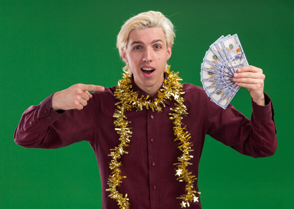圣诞节年轻的金发男子戴着眼镜 脖子上戴着金箔花环 拿着钱指着绿色墙上的隔离墙脖子指尖钱