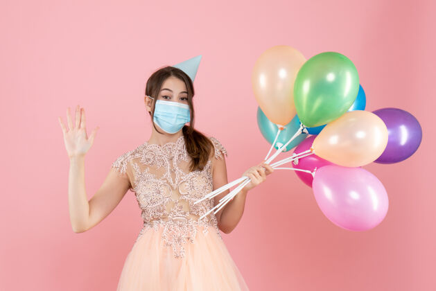医疗戴着派对帽和医用面具的快乐女孩拿着粉色的彩色气球向你问好气球庆祝快乐女孩