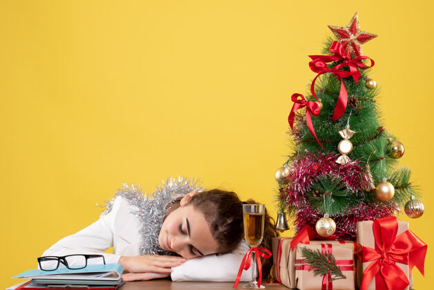微笑正面图：女医生坐在桌子后面 睡在黄色背景上 还有圣诞树和礼品盒圣诞节快乐医生