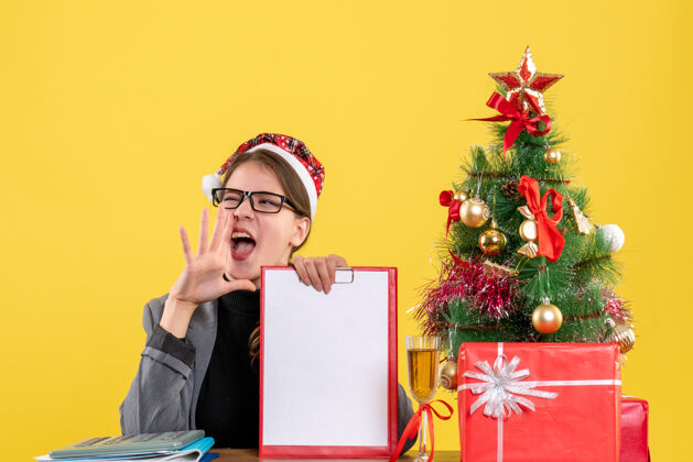 桌子前视图戴圣诞帽的年轻女孩坐在桌旁高喊圣诞树和礼物鸡尾酒人肖像帽子