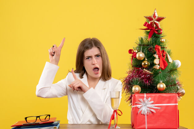快乐正面图：女医生坐在黄色背景的桌子前 放着圣诞树和礼品盒桌子微笑成人