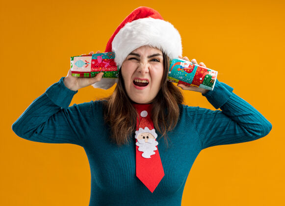 橙色恼怒的成年白人妇女戴着圣诞帽 打着圣诞领带 拿着纸杯 靠近耳朵 橙色背景 留着复印空间圣诞节关闭举行
