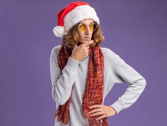 年轻戴着圣诞老人帽 戴着黄色眼镜 脖子上围着暖和围巾的年轻人 带着怀疑的表情看着摄像机 站在紫色背景下思考相机戴着思考
