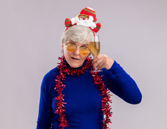 自信自信的老妇人戴着太阳眼镜 戴着圣诞老人的头带 脖子上戴着花环 手里拿着一杯香槟酒 背景是白色的 有复印空间圣诞快乐周围花环