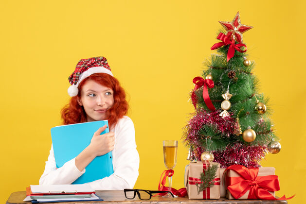 礼物正面图：女医生坐在桌子后面 手里拿着文件 黄色的是圣诞树和礼盒庆祝桌子文件