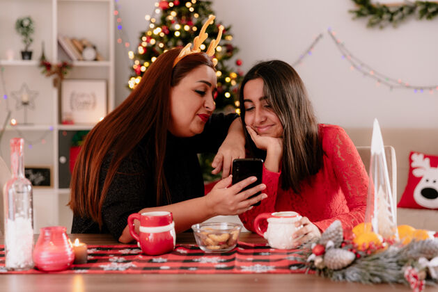 圣诞节高兴的母亲戴着驯鹿的头巾 女儿看着坐在桌边的电话 在家里享受圣诞节的时光高兴享受女儿