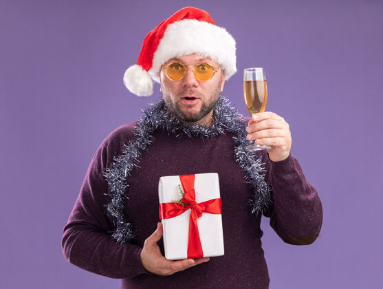 穿着令人印象深刻的中年男子戴着圣诞帽 脖子上戴着金箔花环 戴着眼镜 手里拿着礼包 紫色墙上隔着一杯香槟金属丝脖子花环