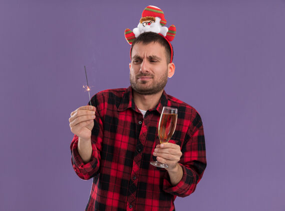 圣诞老人困惑的年轻白种人戴着圣诞老人的头带拿着节日的火花和香槟看火花孤立在紫色墙上的复制空间男人困惑香槟