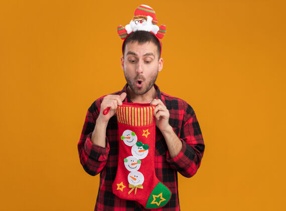 好奇好奇的白人年轻人戴着圣诞老人的头带 手里拿着圣诞长袜 打开里面看 橙色的墙上有一个单独的复制空间长袜圣诞老人里面