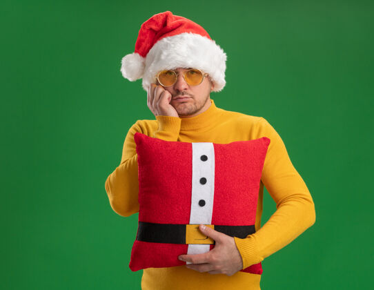 圣诞老人一个穿着黄色高领毛衣 戴着圣诞帽 戴着眼镜 拿着红色滑稽枕头 脸上带着悲伤表情的年轻人站在绿色的墙上站立男人高领毛衣