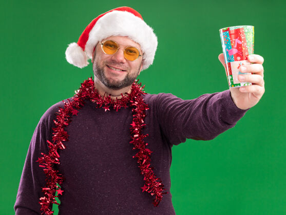圣诞老人微笑着的中年男子戴着圣诞帽 脖子上戴着金属丝花环 戴着眼镜 伸出塑料圣诞杯 孤立在绿色的墙上塑料脖子圣诞节