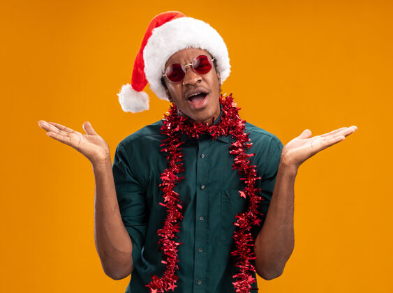 肩膀戴着圣诞帽戴着花环戴着眼镜的非裔美国人困惑地耸耸肩站在橙色的墙上花环站着圣诞老人