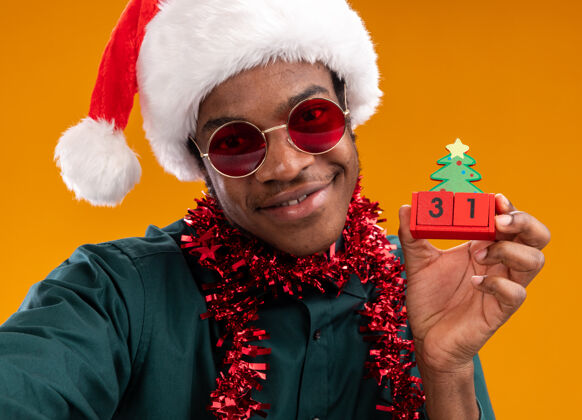 戴着快乐的非洲裔美国人戴着圣诞帽 戴着花环 戴着太阳镜 抱着新年礼物的玩具立方体 微笑着站在橙色的墙上圣诞节帽子花环