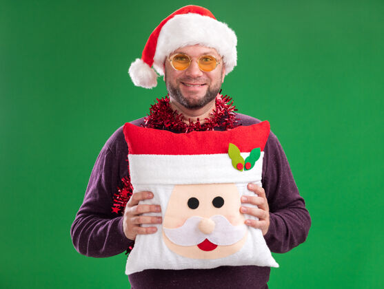 枕头微笑的中年男子戴着圣诞帽 脖子上戴着金箔花环 戴着眼镜 把圣诞老人的枕头孤立在绿色的墙上周围圣诞老人圣诞节