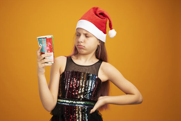 帽子小女孩穿着闪亮的派对礼服 戴着圣诞帽 手里拿着两个五颜六色的纸杯 站在橘色的墙上困惑地看着它迷糊五颜六色杯子