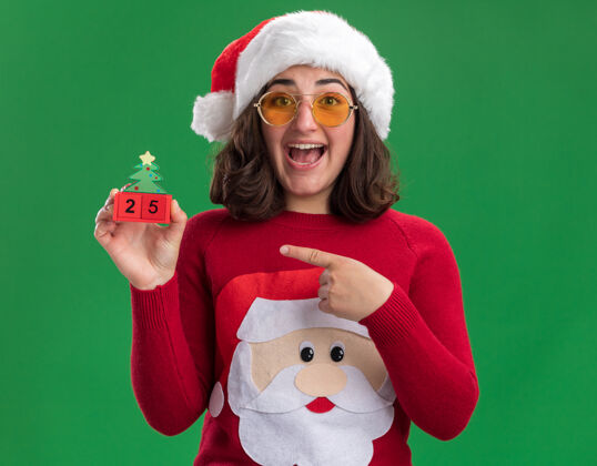 帽子穿着圣诞毛衣 戴着圣诞帽 戴着眼镜 手里拿着25号玩具方块 食指指着玩具方块 站在绿色的墙上开心地笑着穿着立方体手指