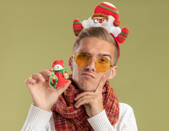 橄榄可疑的年轻帅哥戴着圣诞老人的头带和围巾 手里拿着雪人的圣诞饰品 手指贴在脸上 孤零零地贴在橄榄绿的墙上雪人围巾圣诞老人