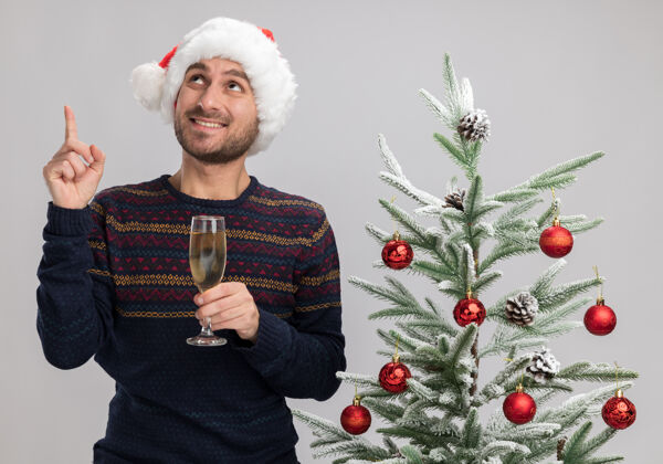 微笑带着圣诞帽的年轻白人微笑着站在圣诞树旁 手里拿着一杯香槟 望着白色背景上与世隔绝的人群年轻圣诞指着