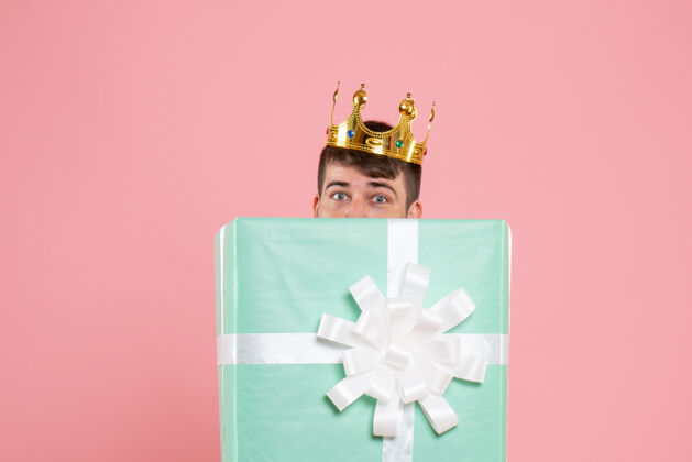 年轻年轻人在礼物盒内的正面视图 粉红色墙上有皇冠年轻男性聚会皇冠