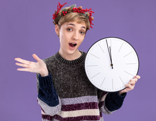 空虚令人印象深刻的年轻漂亮女孩戴着圣诞花环手持时钟显示空手孤立在紫色的墙上印象深刻展示头