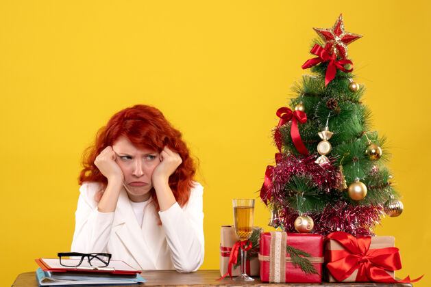 办公室女工人坐在桌子后面 手里拿着圣诞礼物和黄色的圣诞树桌子女工情感