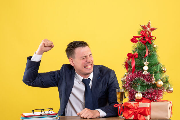 男人愤怒的男人举起手坐在圣诞树旁的桌子前 黄色的墙上挂着礼物公司商务愤怒的男人