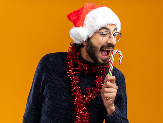 花环兴奋的年轻帅哥戴着圣诞帽 脖子上戴着花环 手里拿着圣诞糖果 孤零零地站在橙色的墙上圣诞节帽子兴奋